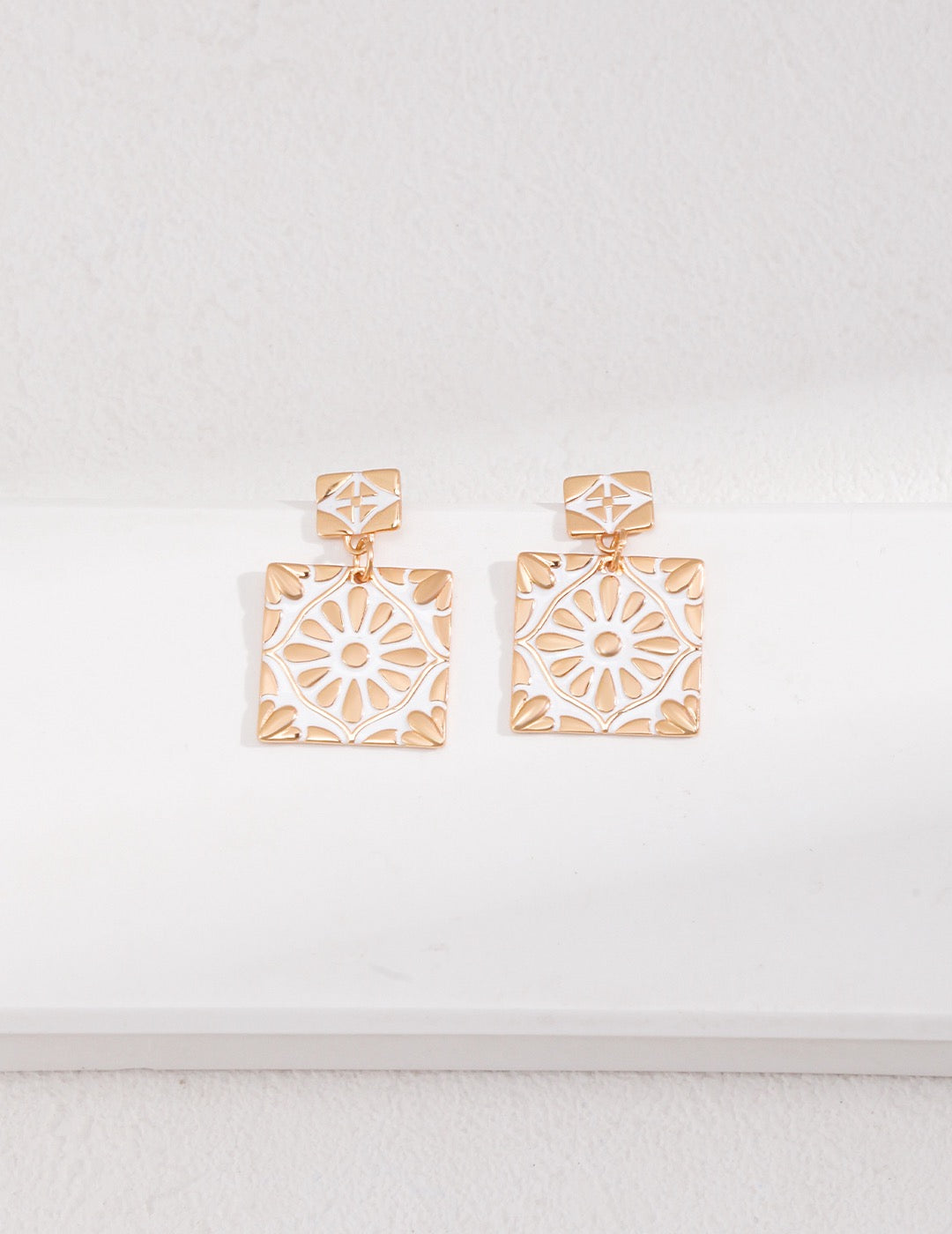 Exquisite drop studs with a vibrant white enamel inlay, set in a geometric square with a delicate floral design, crafted in gold.